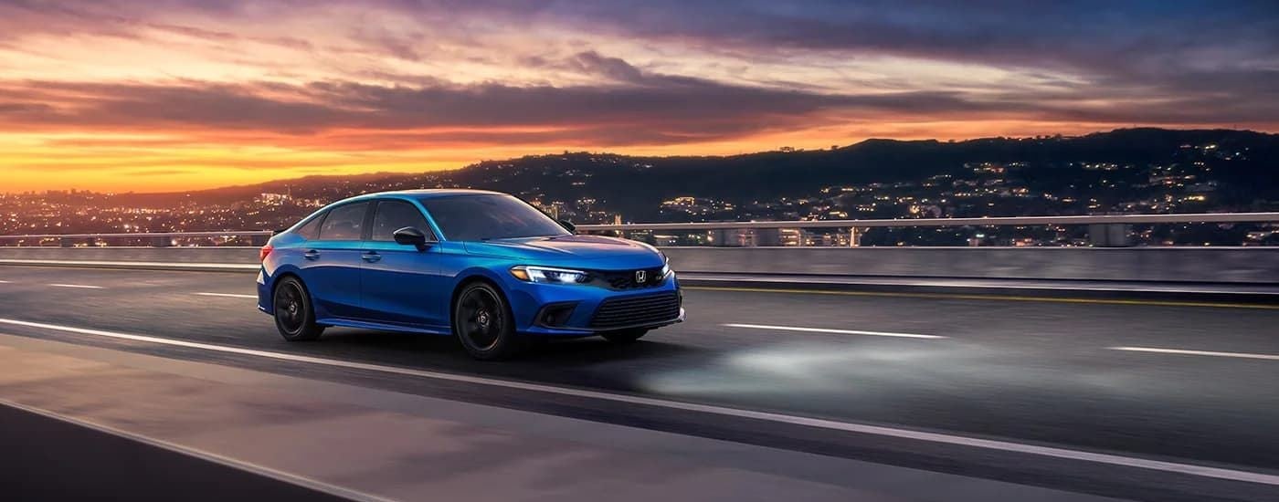 A blue 2022 Honda Civic Si is shown driving past a distant city at sunset.