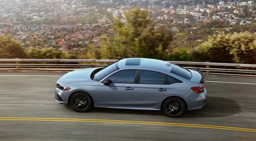 A grey 2022 Honda Civic Si is shown from the side driving on a highway overlooking a city.