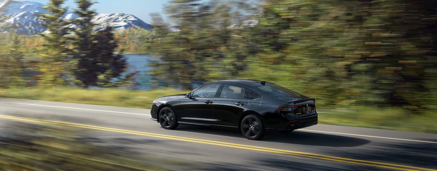 A black 2023 Honda Sport-L is shown from the side while driving on a road next to a lake.