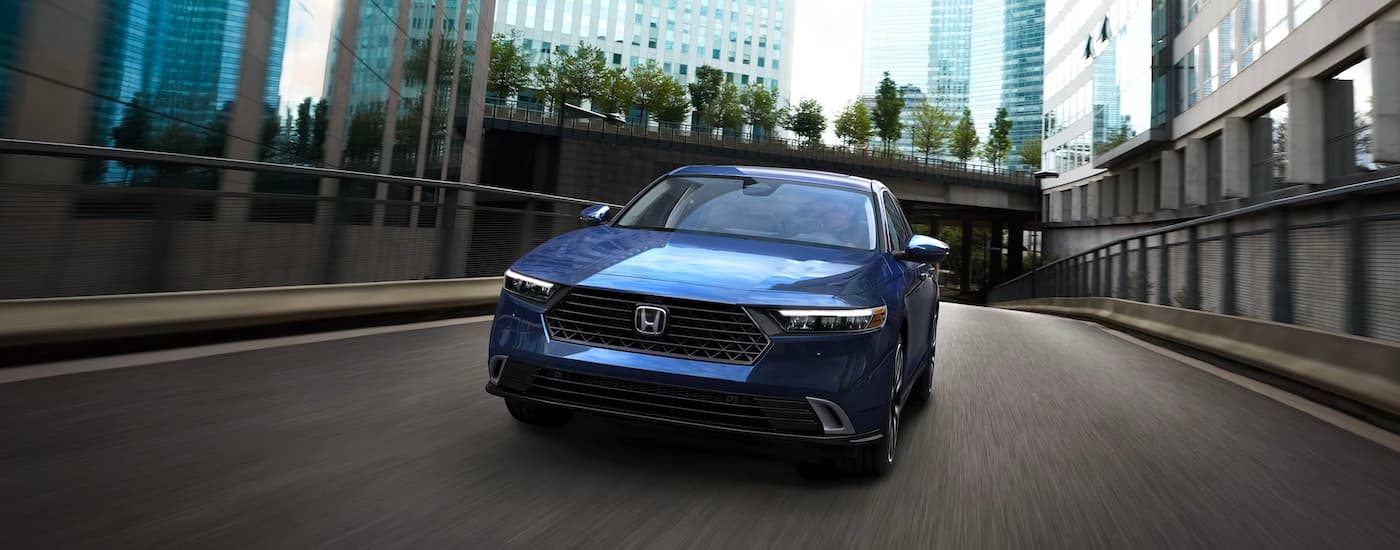 A blue 2023 Honda Accord Sport Hybrid is shown from the front driving on a city highway.
