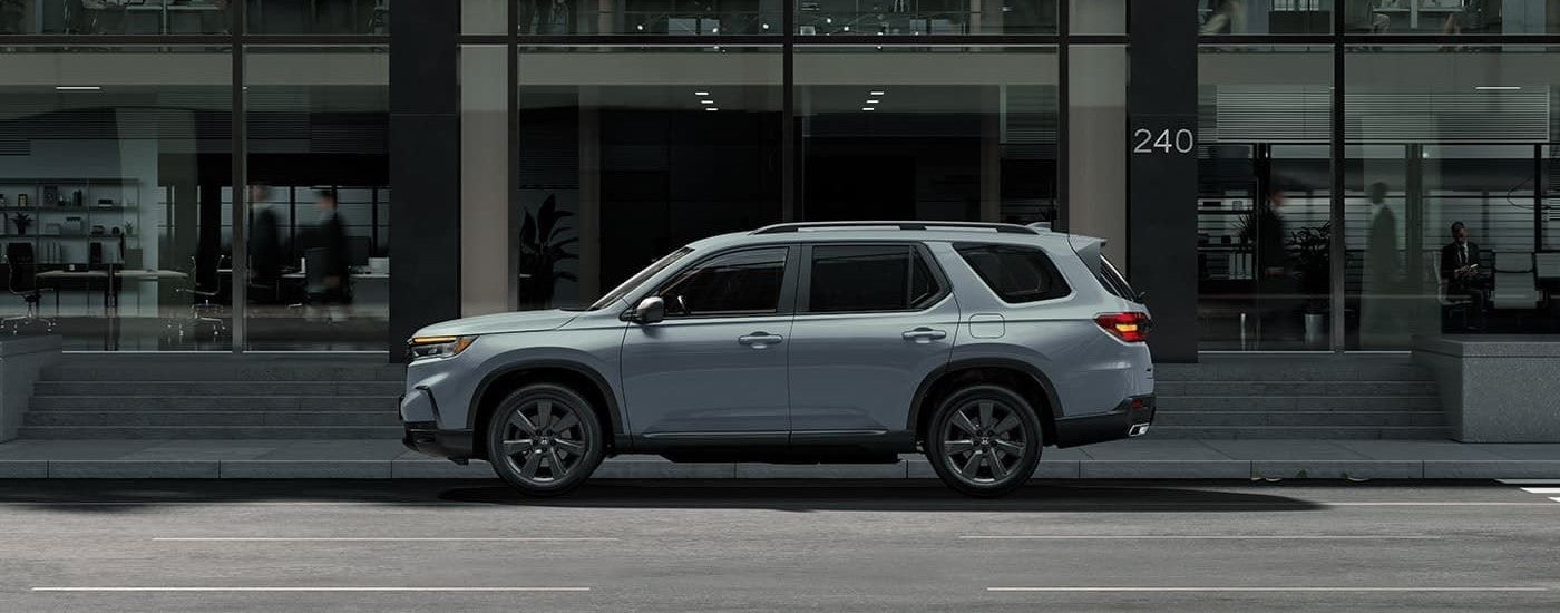 A grey 2023 Honda Pilot Sport is shown from the side part on the edge of a city street in front of a metal and glass building.