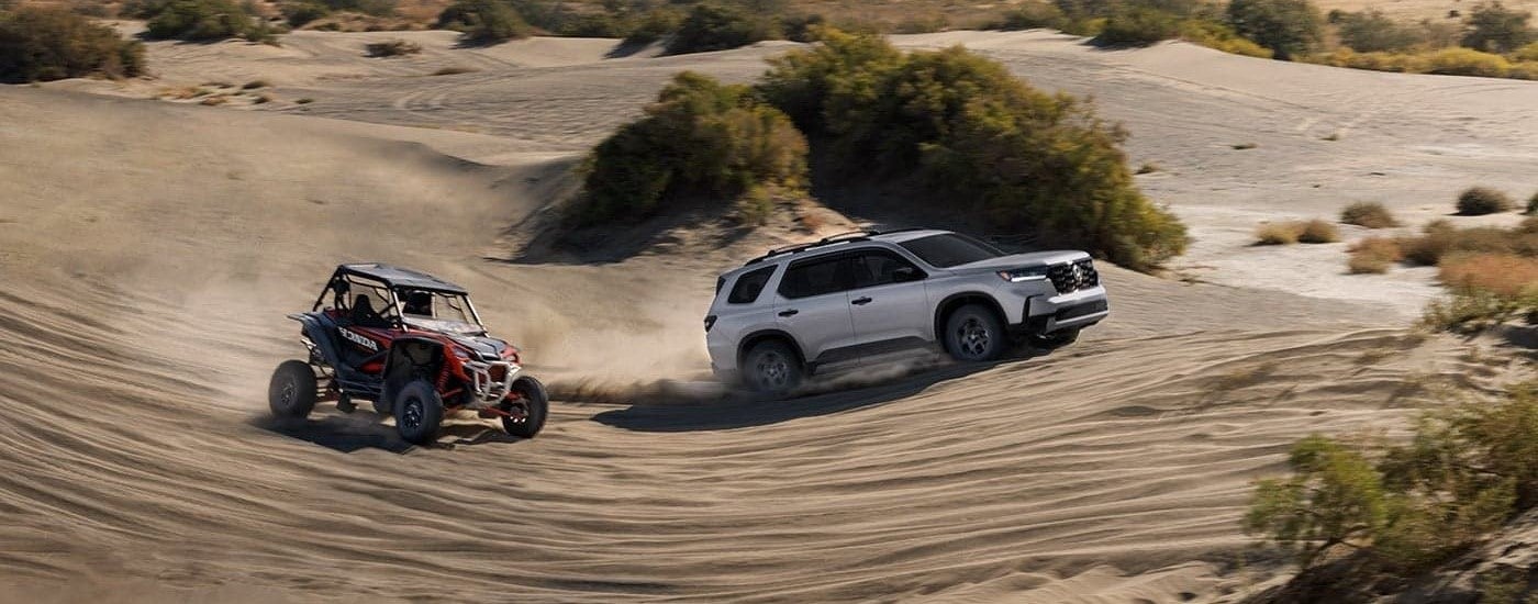 A silver 2023 Honda Pilot Trailsport is shown off-roading on a sandy area next to a red UTV.