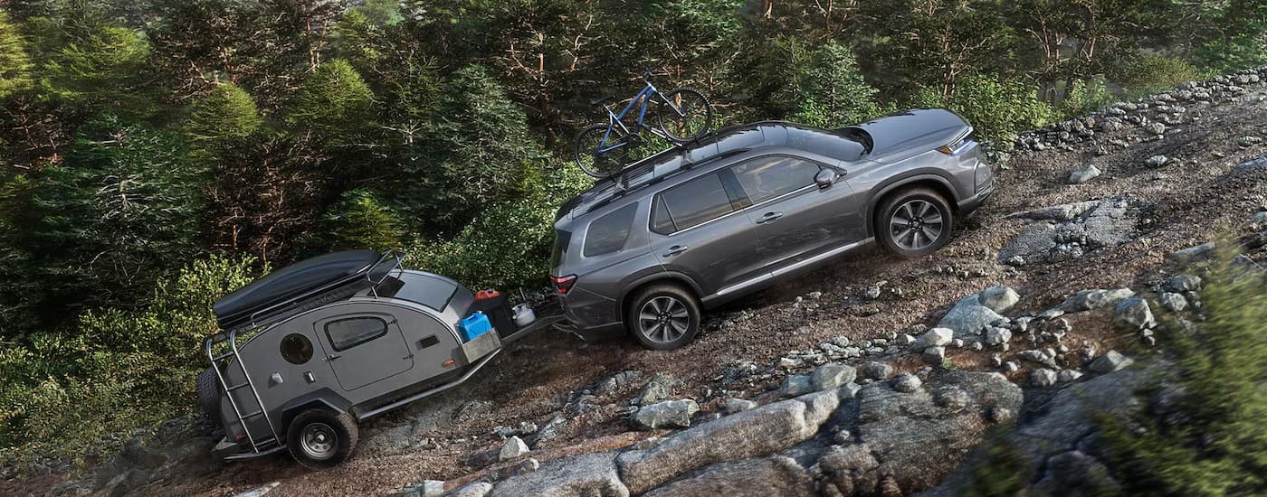 A grey 2025 Honda Pilot is towing a small grey camper up a rocky hill.