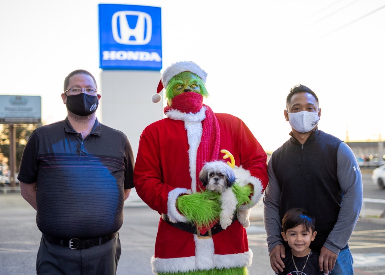 Grinch in a Santa Suit Holding a Puppy Next to Two Men and a Boy in Front of Honda Sign